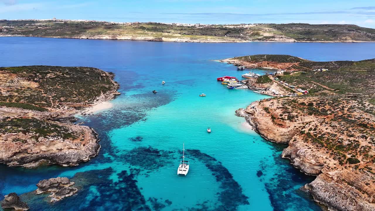Vista aérea da Lagoa Azul em Comino, Malta, com vários barcos ancorados em águas claras e turquesa entre costas rochosas e pequenas ilhas sob a luz brilhante do dia