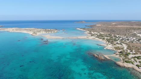 Vista-De-Drones-En-Grecia-Volando-Sobre-La-Playa-Estrecha-De-Arena-Blanca-De-Elafonisi,-Agua-Azul-Clara-A-Los-Lados-Y-Paisaje-Marrón-En-Un-Día-Soleado