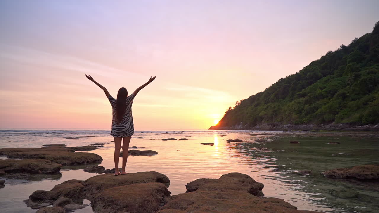 una mujer de pie sobre una roca en una playa tropical y levantando las manos a la luz del sol al atardecer