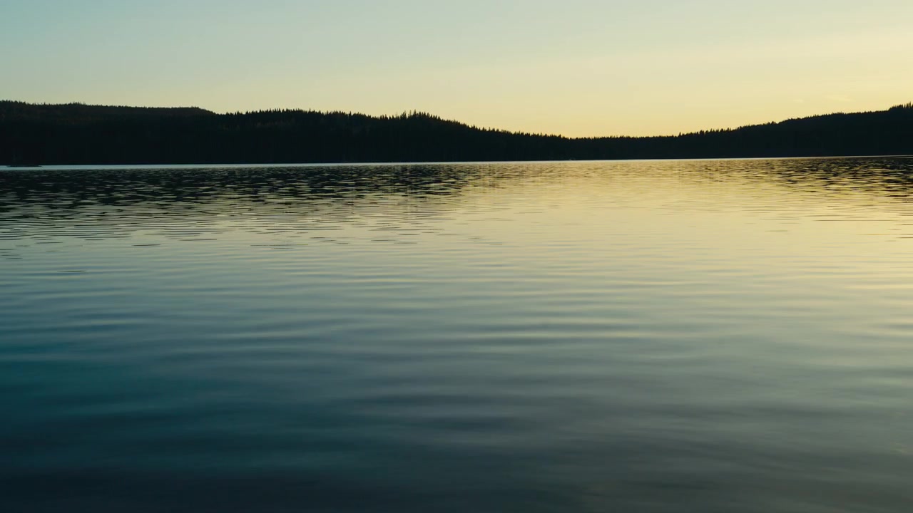Blide krusninger bevæger sig hen over en rolig sø i skumringen, mens en mørk skovhorisont og falmende aftenlys skaber et fredfyldt og minimalistisk nordisk vandlandskab.