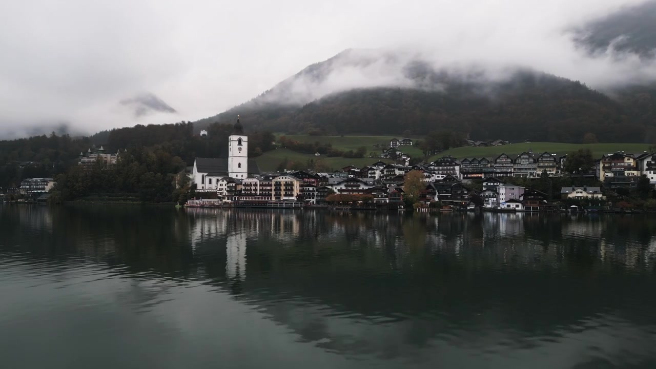 Bevægelig luftdroneoptagelse af Wolfgang am Wolfgangsee langvejsfra på en regnvejrsdag tæt på søen med skyer på bjergene.