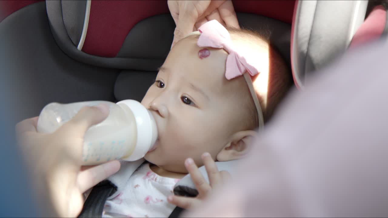 귀여운 아시아 아기 소녀가 차 좌석에서 병에서 차분하게 우유를 마시고 어린이의 승객 안전 제품과 도로 여행의 필수품 안전하고 편안한 여행과 신생아 개념의 자동차 여행.