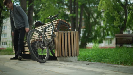 young boy in glasses rides bicycle into peaceful park, parks it between wooden bench and nearby structure, rests bike on bench, then sits down to relax, leaning back, surrounded by lush greenery