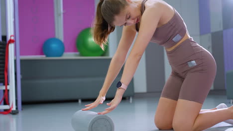 lady in brown outfit performs flexibility exercises on a mat, stretching forward over a foam roller, fitness ball visible in background of well-equipped gym