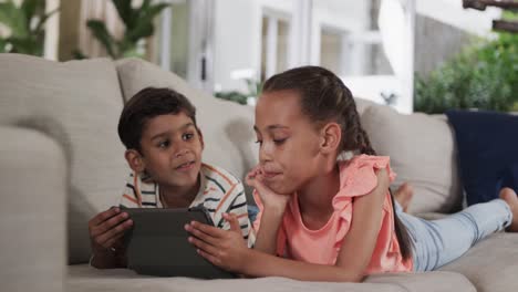 feliz hermano y hermana biracial acostados en el sofá y usando la tableta en casa, cámara lenta