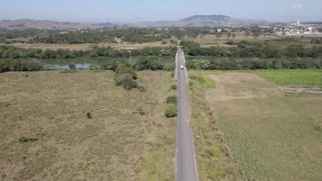 Camión-Solitario-En-La-Carretera-Vista-Aérea-Desde-Lejos-Cruzando-Un-Puente-Hacia-Un-Pequeño-Pueblo-En-Un-Día-Soleado