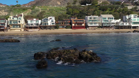 4K-Aerial-drone-shot-of-Beach-coastline-with-the-blue-Pacific-Ocean-with-waves-coming-in-and-beach-with-nice-houses-on-the-background