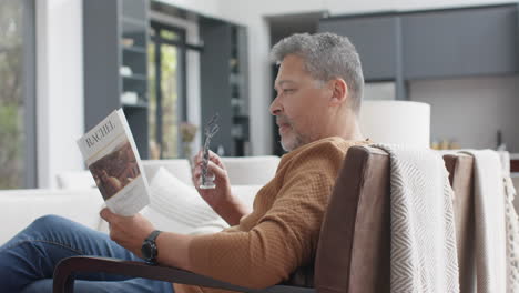 Hombre-Birracial-Senior-Enfocado-Sentado-En-Un-Sillón-Leyendo-Un-Libro-En-La-Sala-De-Estar,-Cámara-Lenta