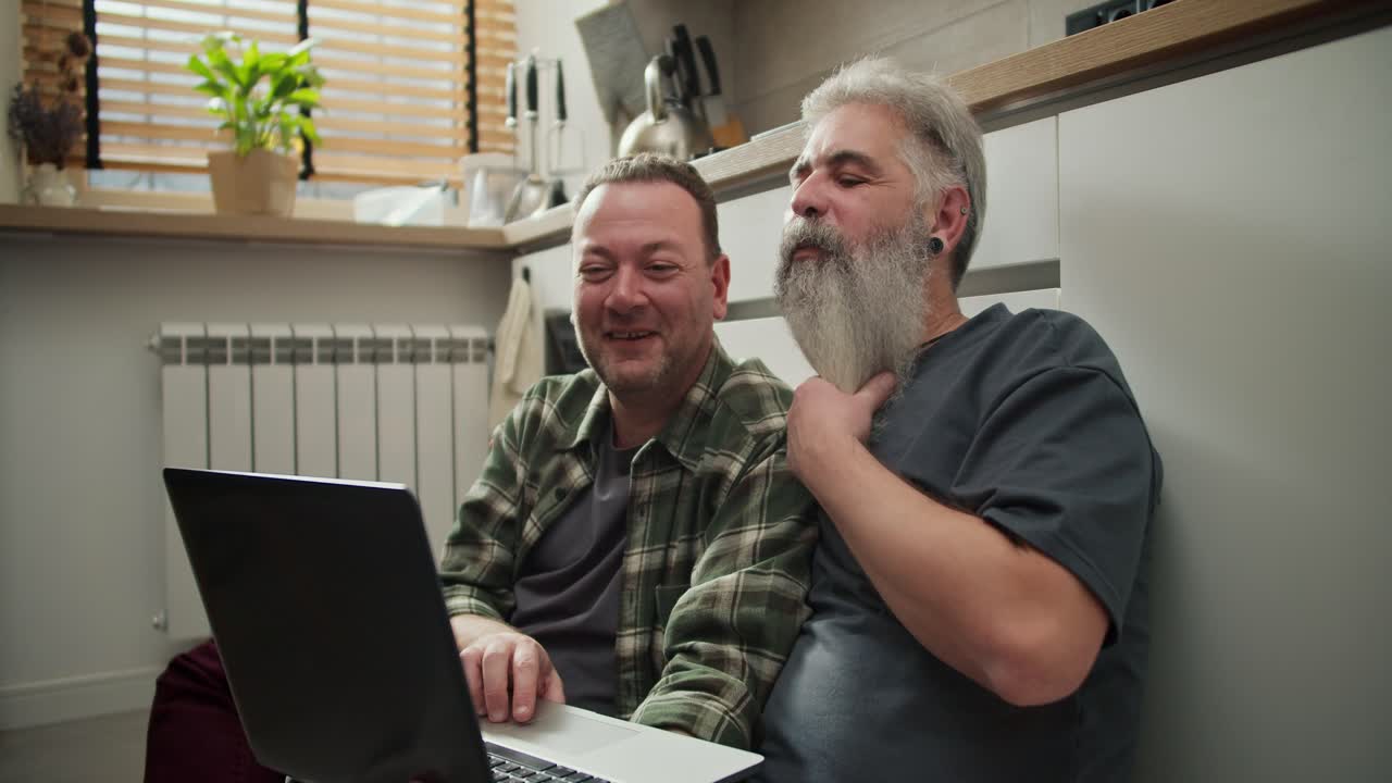 A happy elderly man with gray hair and a lush gray beard straightens his beard while sitting and watching a funny video on a laptop with his brunette boyfriend with stubble in a Green checkered shirt sitting on the floor in the kitchen