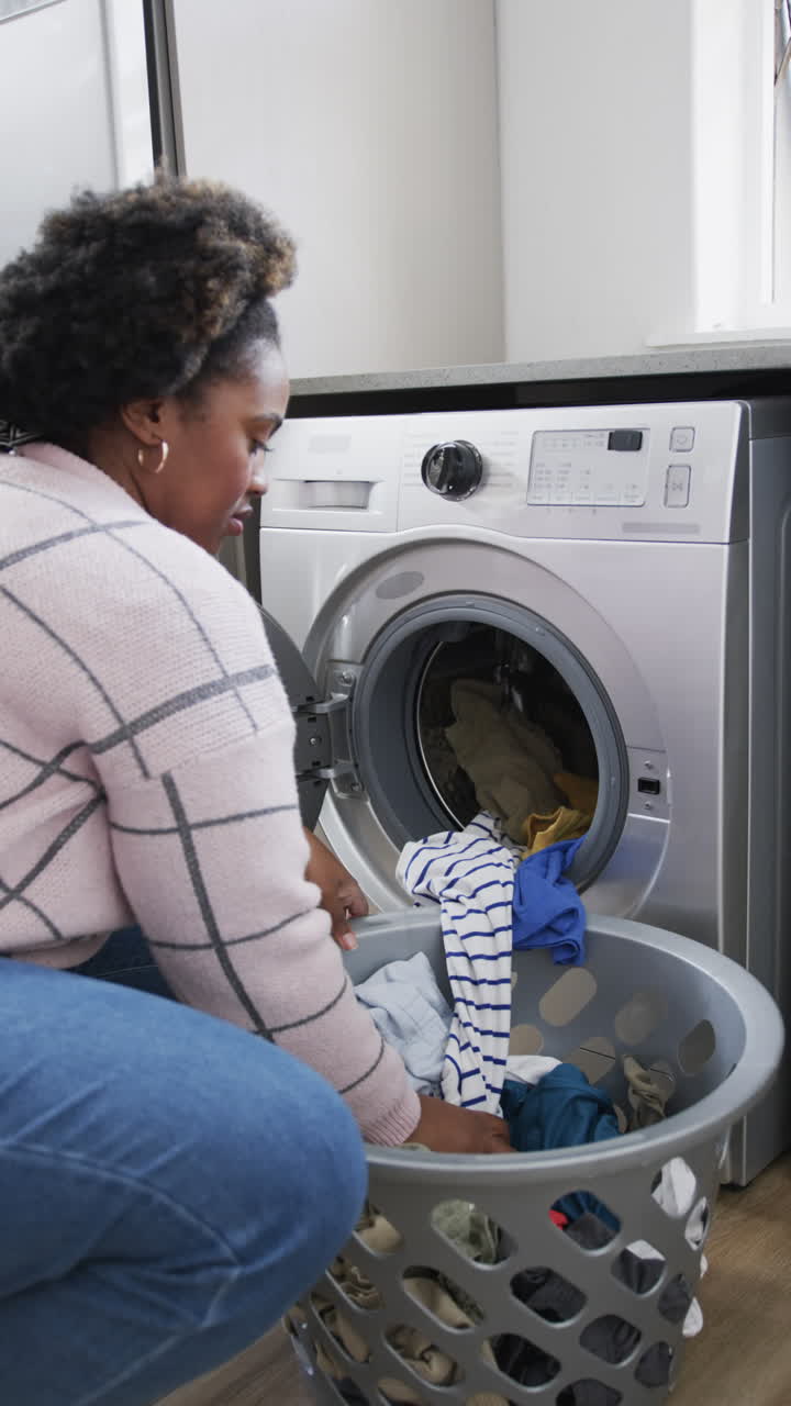 Vertical video of african american woman using washing machine and doing laundry at home