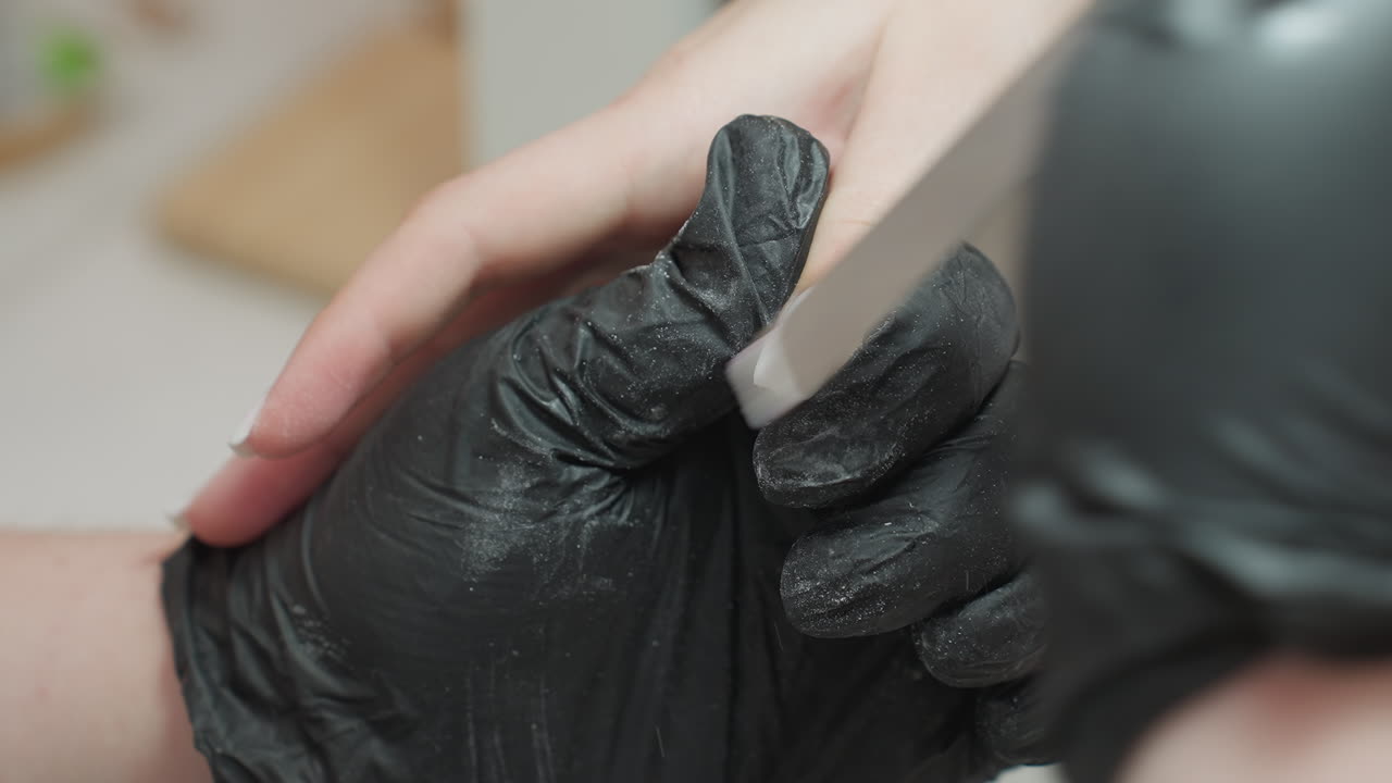 Close-up view of nail technician wearing black glove carefully filing client thumb using manual nail file during manicure session. Focus on precision, cleanliness, and professional hand grooming