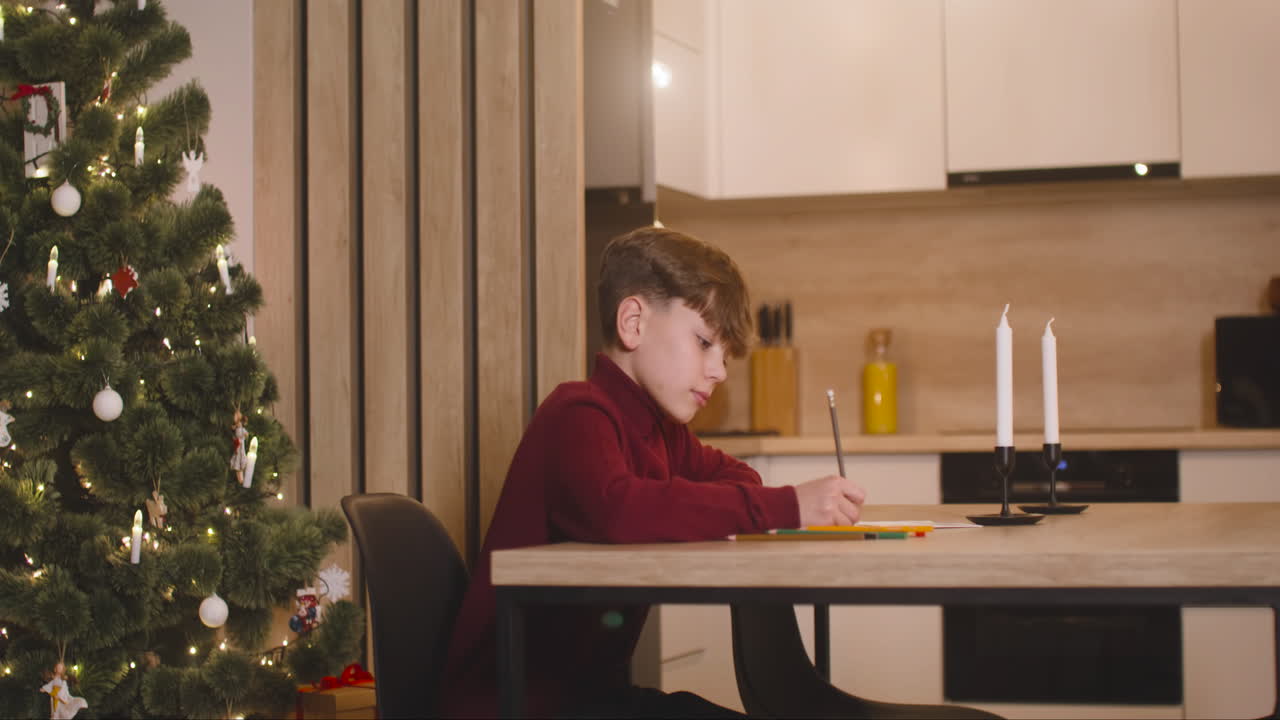 vista lateral de un niño con cuello alto rojo escribiendo una carta de deseos sentado en una mesa en una habitación decorada con un árbol de navidad