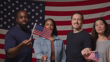 studio portrait shot of multi-cultural group of friends waving miniature stars and stripes flags in front of american flag celebrating 4th july independence day party