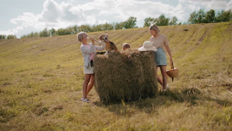 woman holding dog paw atop a haystack while another woman carries a picnic basket, straw hat rests on the hay, and a child peeks from behind, sunlit countryside