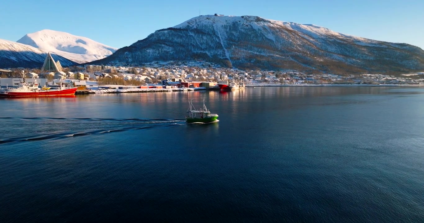 Fischerboot fährt unter der Tromsø-Brücke hindurch, mit der Stadt im Winterschnee. Luftaufnahme.