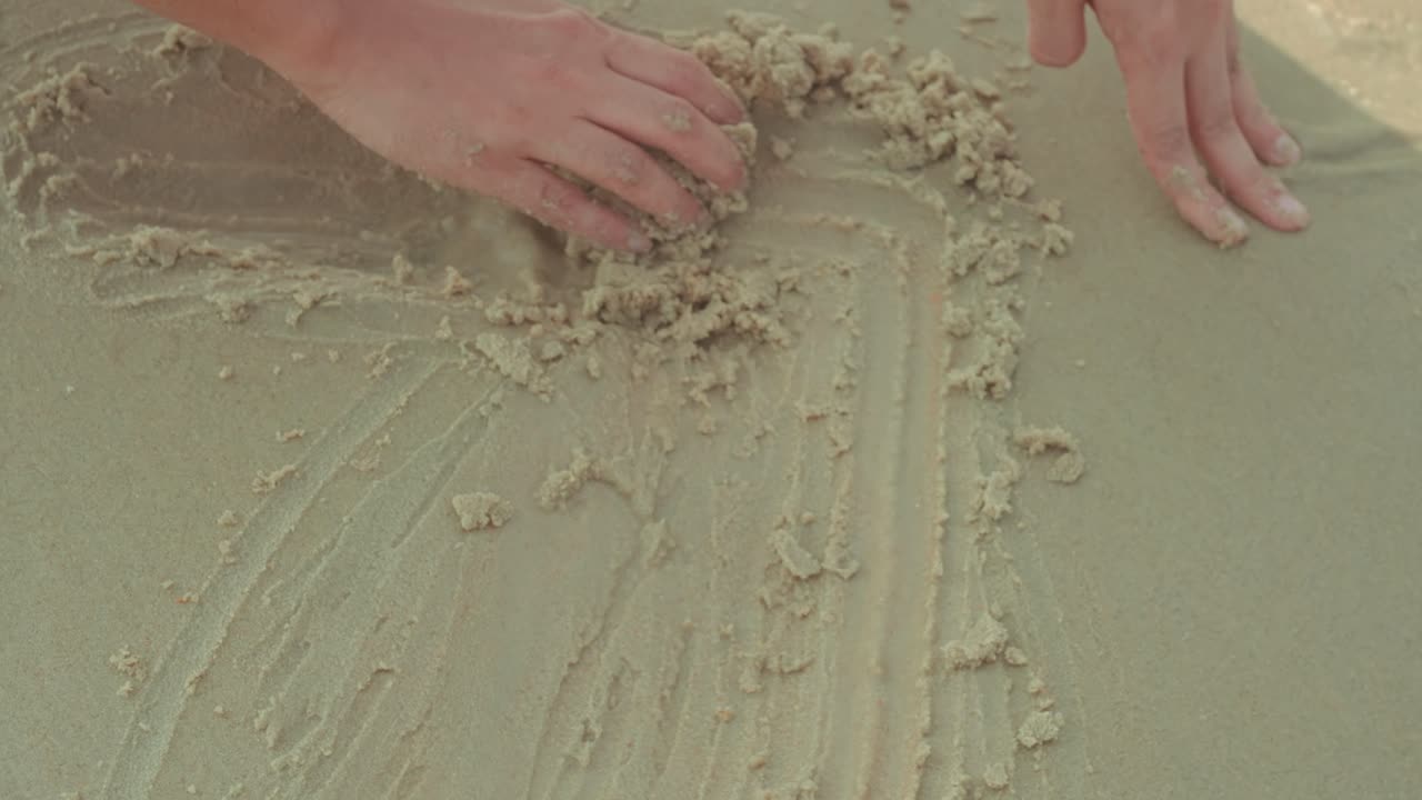 Primer plano de una mano dibujando líneas y formas en la suave arena de la playa. Momento creativo minimalista con textura natural, ideal para conceptos de infancia, creatividad y ocio.