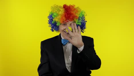 Senior-old-woman-clown-in-wig-waves-her-hands,-smiling.-Yellow-background