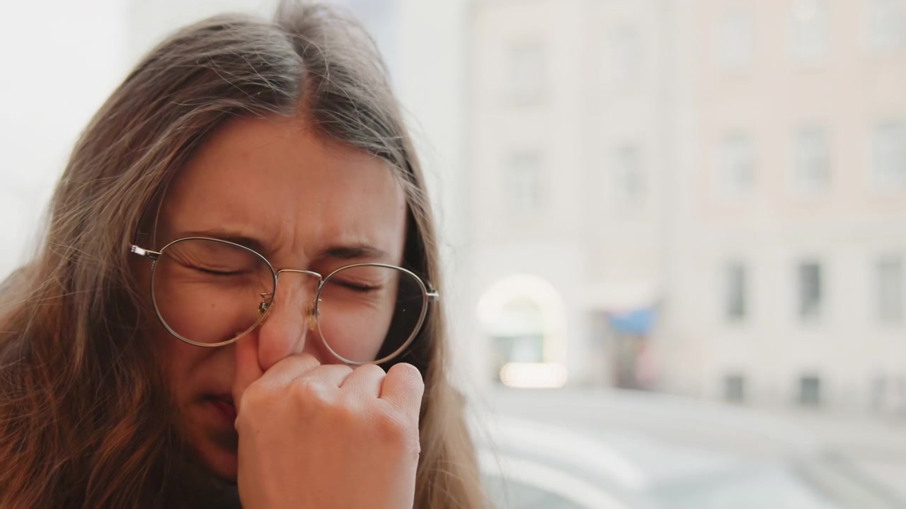 Mulher cobrindo o rosto enquanto espirra ao ar livre. Incidente súbito de espirro capturado com olhos lacrimejantes e expressão de desconforto. Momento espontâneo de uma mulher espirrando numa rua fria durante o seu trajeto.