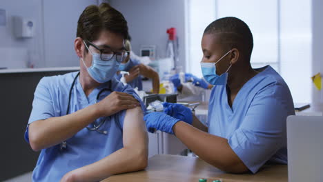 African-american-female-doctor-giving-covid-vaccination-to-asian-male-colleague,-both-in-face-masks