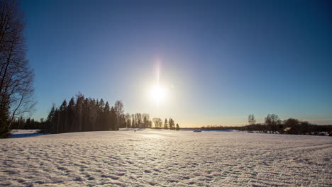 Eine-Untergehende-Sonne-Vor-Blauem-Himmel-Mit-Kleinen-Weißen-Wolken,-Die-über-Ein-Verschneites-Winterfeld-Mit-Bäumen-Im-Hintergrund-Ziehen