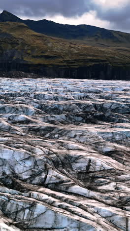 ridges of blue and black ice covering the valley among the mountains. top view on the ancient glacier of iceland under the dramatic sky. vertical video.