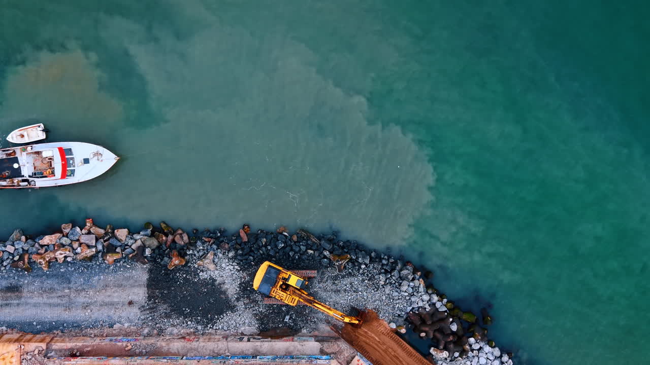 Vista dall'alto di un escavatore che sposta sabbia su un molo roccioso. Escavatore giallo che lavora con terra e rocce al bordo dell'acqua di mare turchese
