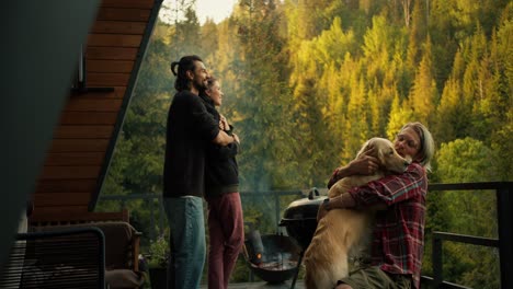 un grupo de amigos en un picnic con vistas a las montañas y el bosque de coníferas. un chico moreno está abrazando a una chica morena, y un hombre rubio está acariciando a un perro grande de color claro