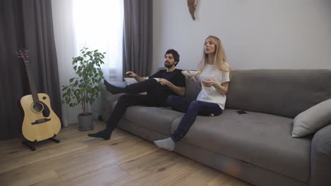 Happy-couple-eating-dinner-from-white-bowls,-sitting-on-the-sofa-and-turn-on-TV