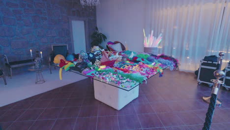 table filled with various colorful party decorations, including hats, masks, feather boas, and glowing accessories, set in a room with stone walls and ambient lighting