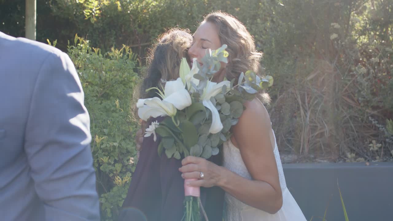 Bride and female guest greeting, reaching out and hugging, bride cradling large bouquet by hillside