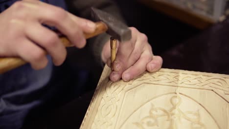 A-craftsman-carving-wooden-ornament-on-backgammon-sitting-in-his-working-place.-Close-up-footage-of-a-man-working-on-details-using-small-wooden-stick-and-hammer