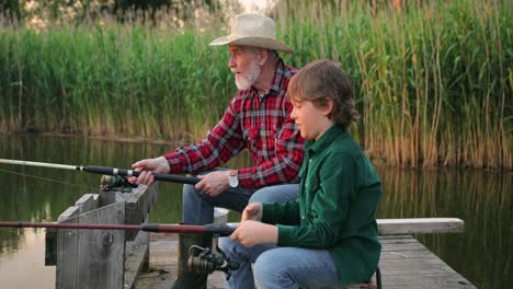 side view of a teen boy sitting with his grandfather on the lake pier fishing together