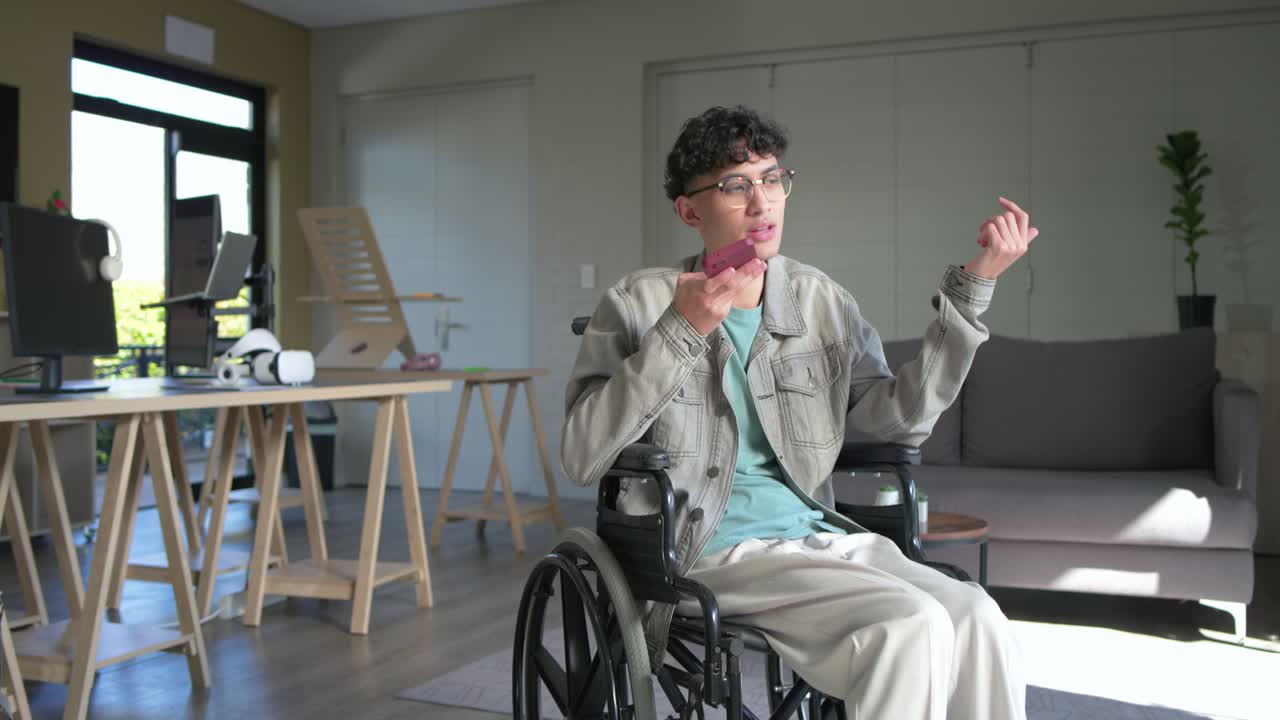 Mid-adult man sitting in wheelchair lifting smartphone to mouth responding to call in office