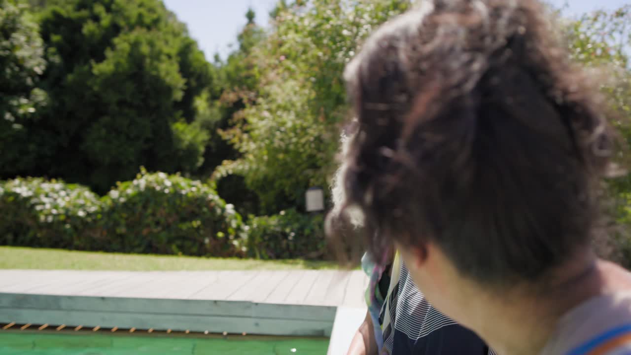 Amiche in piscina nel cortile, un'anziana con i capelli intrecciati conduce la conversazione mentre un'amica sorseggia una bevanda rosa.