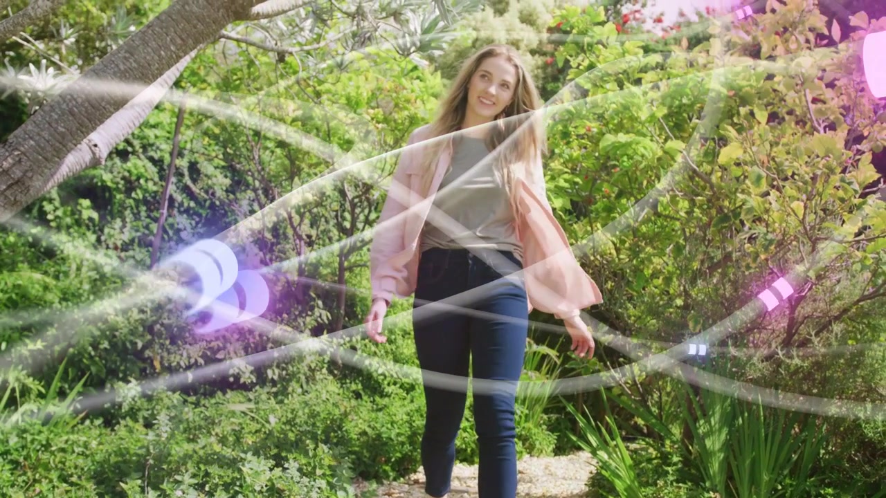 Una donna cammina su un sentiero in giardino verso la telecamera, con nastri e sfere che la avvolgono per un effetto moda.