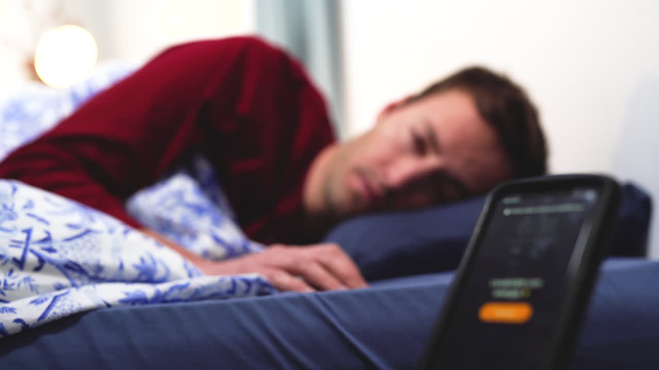 joven caucásico en pijama rojo apague el despertador que suena por la mañana y se despierte de una cama cómoda listo para comenzar el día