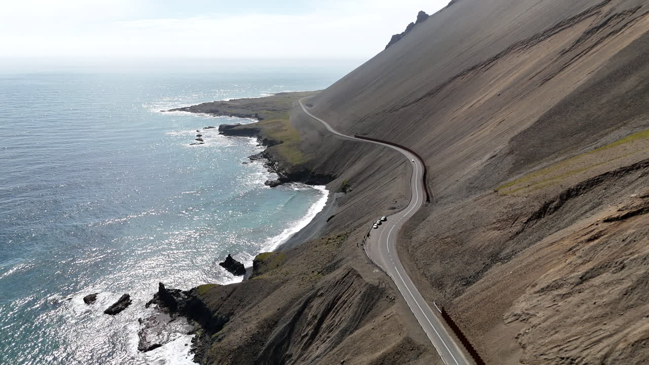 Aerial view of ocean waves hitting the rugged cliffside mountains of Iceland, revealing a winding coastal road, steep rocky slopes, and bright blue water stretching along the dramatic shoreline
