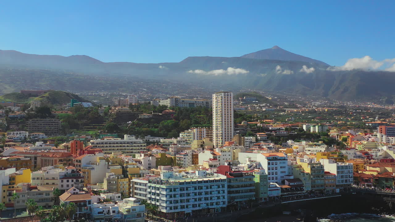 Vista panoramica della città e del porto di Puerto de la Cruz, Isole Canarie, Spagna, con montagne e il Pico del Teide sullo sfondo, sulla costa dell'Oceano Atlantico, mare e cielo blu, ripresa aerea 4K.