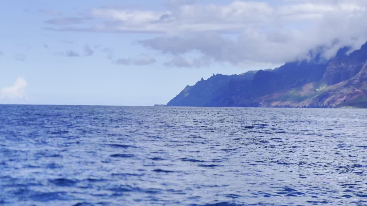 Cinematic long lens panning shot from a boat of the beautiful Na Pali Coast with spinner dolphins leaping out of the ocean in Kaua'i, Hawai'i