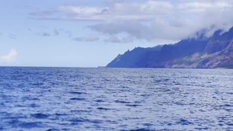 Panorámica-Cinematográfica-Con-Lente-Larga-Desde-Un-Barco-De-La-Hermosa-Costa-De-Na-Pali-Con-Delfines-Saltando-Del-Océano-En-Kaua&#39;i,-Hawai&#39;i