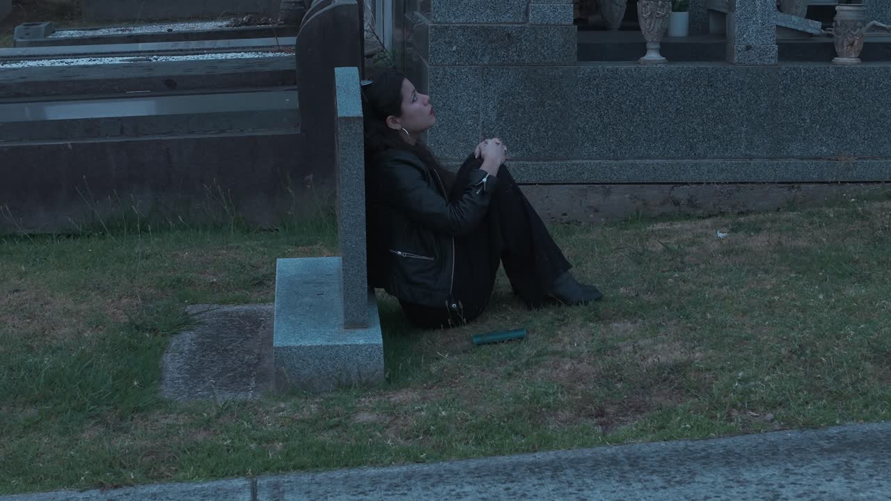 Uma mulher senta-se pensativamente ao lado de uma lápide, envolvida em tons de pôr do sol em um cemitério em Ferntree Gully, Melbourne. O cenário sereno evoca introspecção e melancolia em meio à luz tranquila.