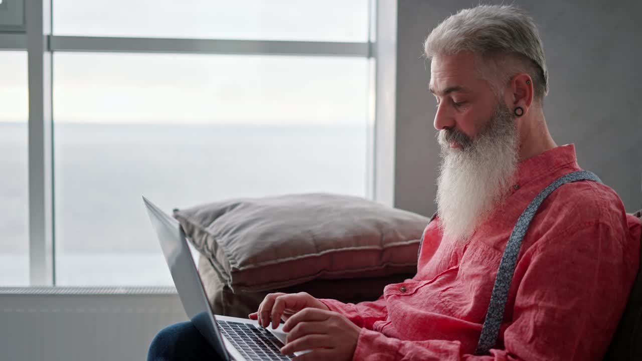 Side view of an elderly man with gray hair and a full beard with a ring in his ear and a pink shirt sitting on a modern sofa and typing on his gray laptop in a modern apartment overlooking the sea