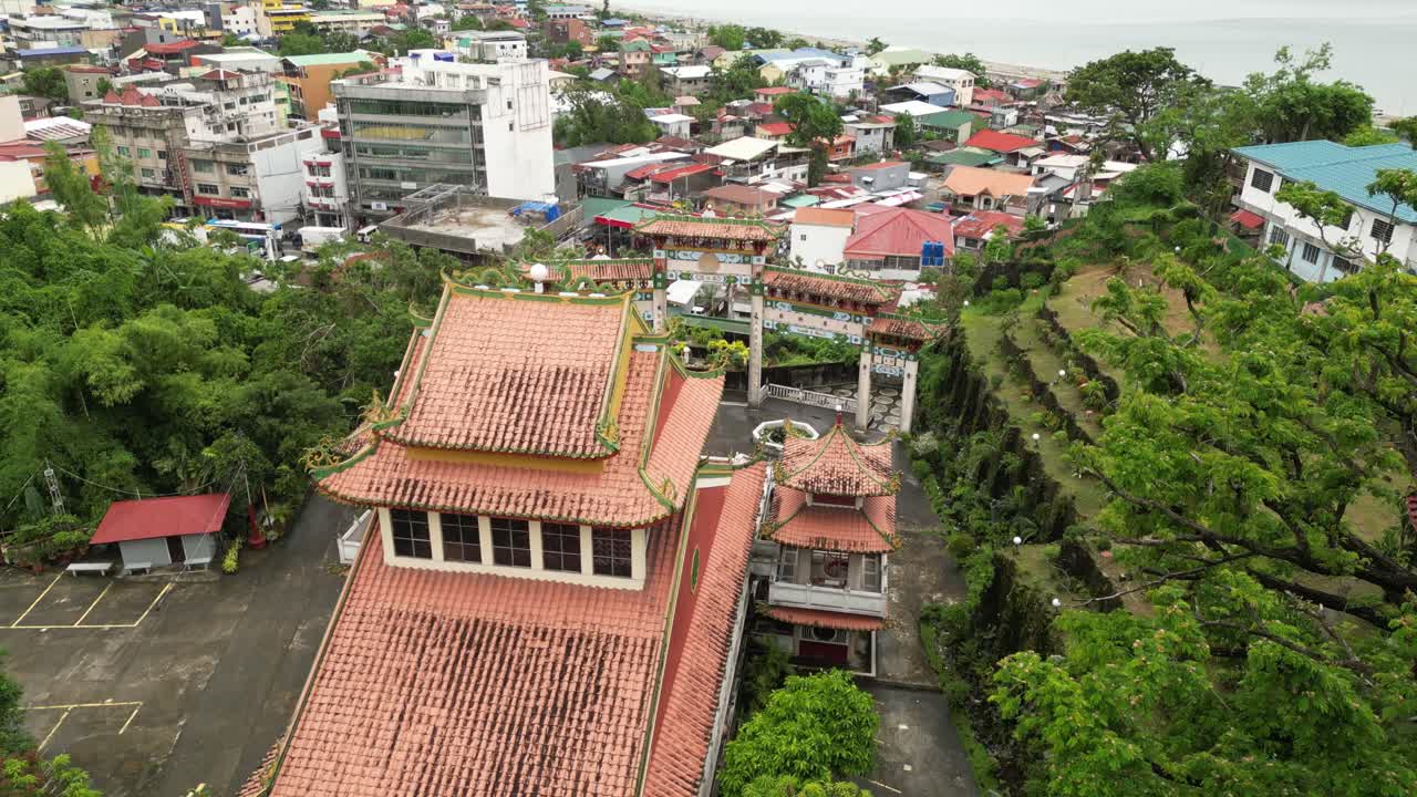 An aerial view opens on a temple complex with ornate red-tiled roofs and a decorative gate. The camera slowly zooms out, revealing a circular garden, surrounding buildings, and lush greenery