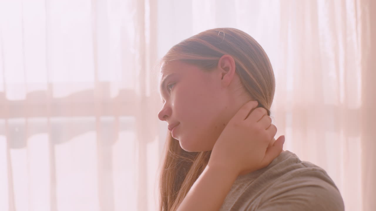 Young woman with long hair gently touches neck muscles while sitting near window with soft curtains, bathed in morning sunlight, expressing discomfort or stretch in calm indoor atmosphere