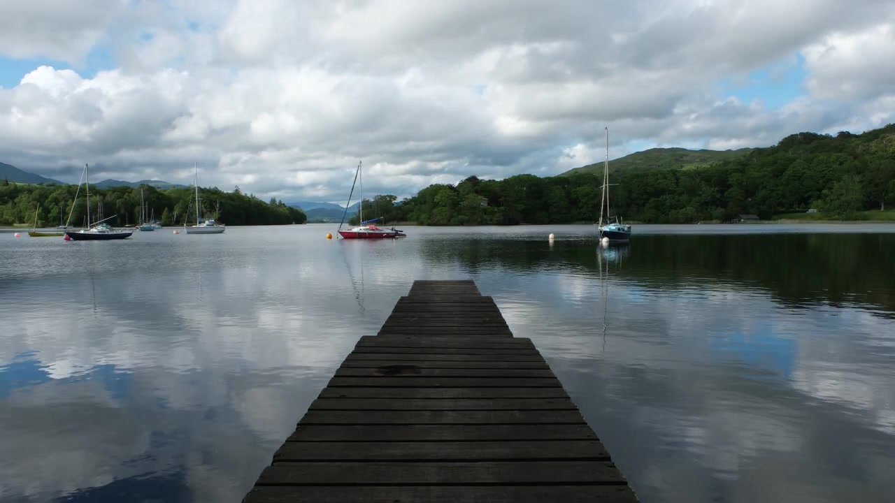 Vista in timelapse da un pontile di legno attraverso il lago Coniston nel Lake District, con barche a vela ormeggiate, nuvole alla deriva, riflessi calmi sull'acqua e boschi e colline circostanti.