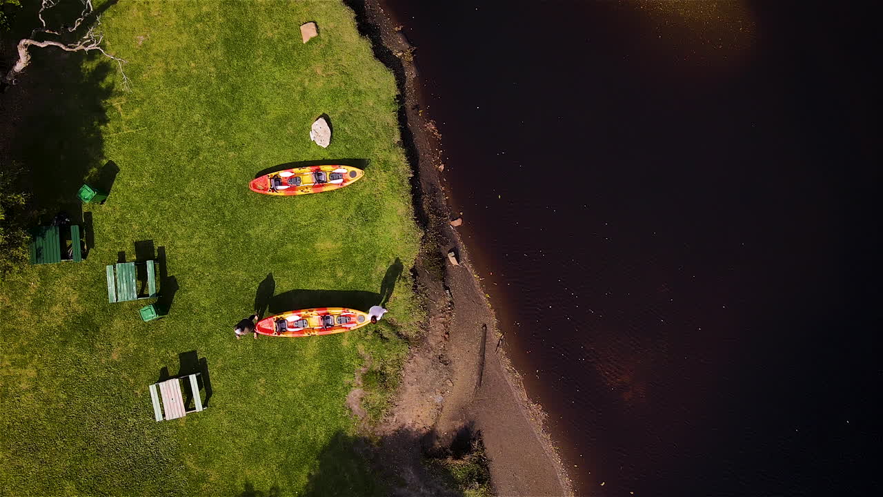 Two women carrying a double kayak to murky river, from grassy river bank, drone