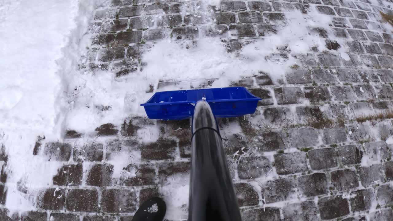 POV pushing a blue shovel through packed slush on a cobblestone driveway, removing footprints and icy chunks to improve traction and reduce slip risk after a cold-weather storm