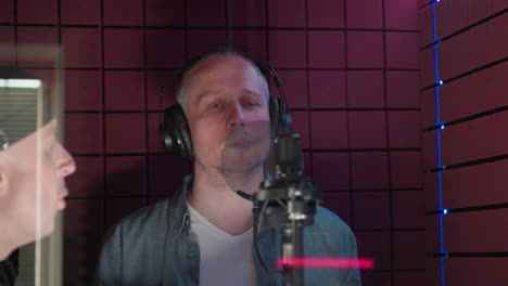 a man in a blue shirt and white inner shirt stands in a red soundproof room, wearing headphones and singing with intense focus. a second man, partially visible in a black cap, observes the session