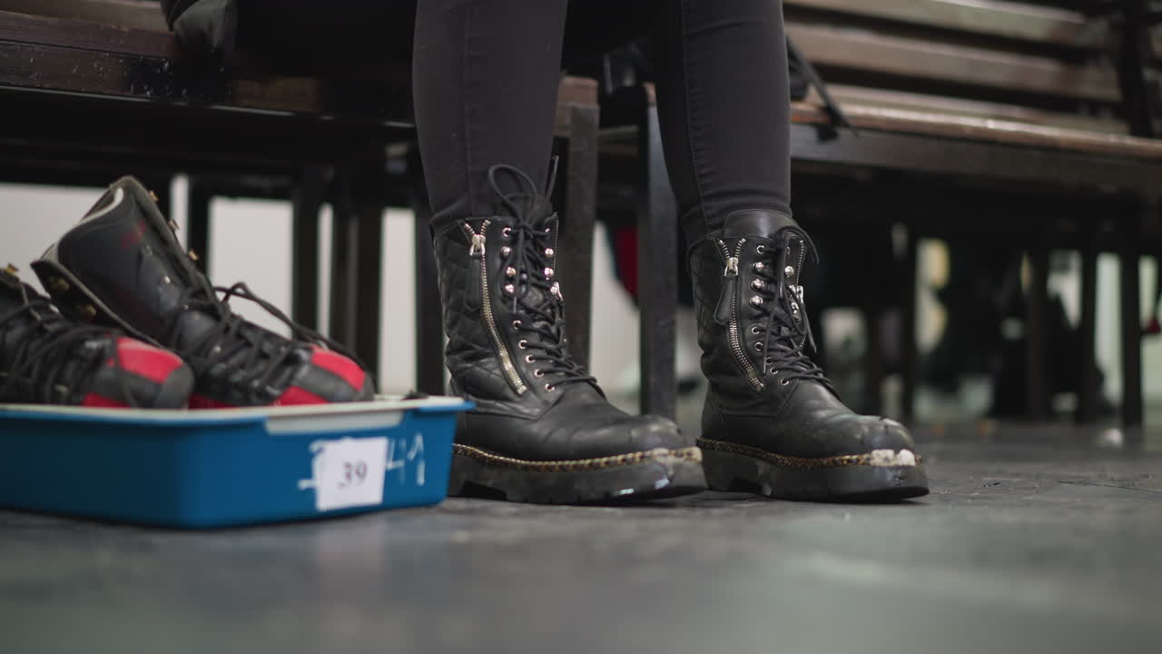 Hand changing shoe by lacing black leather boots with zippers while red and black ice skates rest in blue tray on scratched indoor floor preparation for skating activity winter transition moment