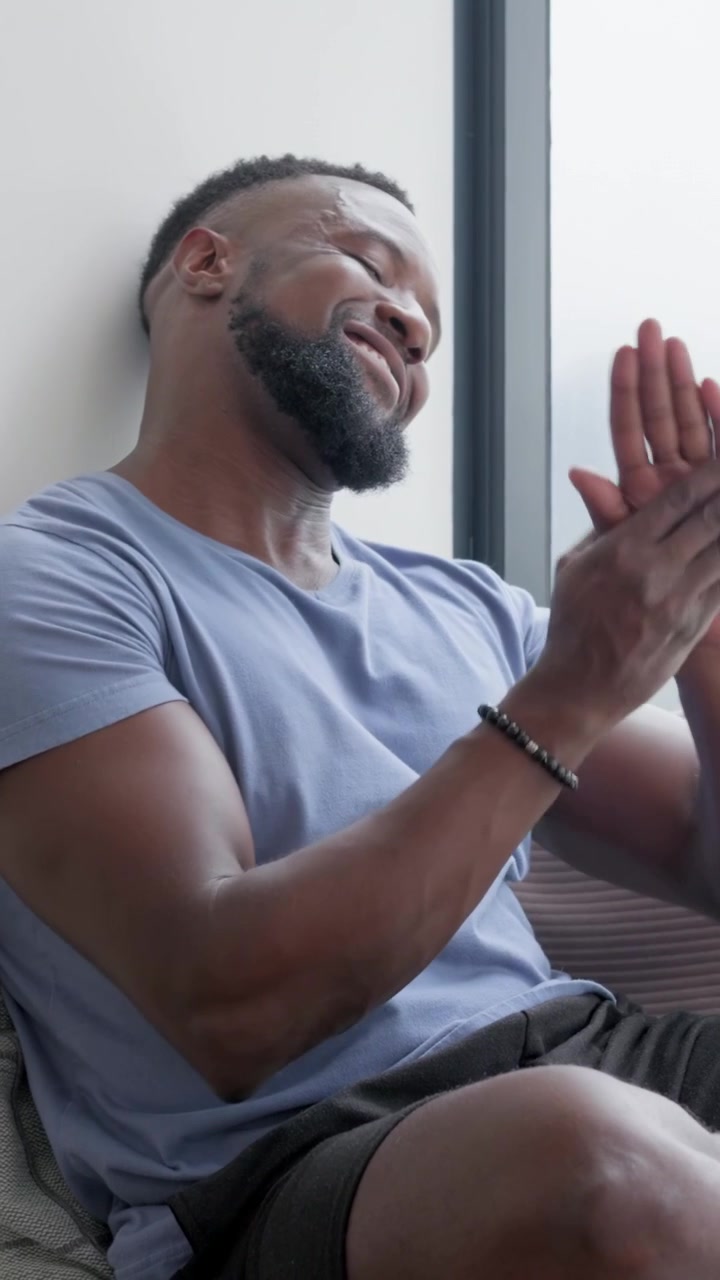 Vertical video: Noticing beaded bracelet, man in blue tee adjusting it on couch near window